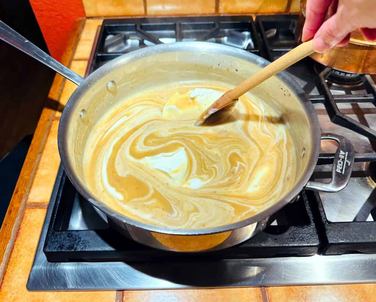 Creme fraiche being swirled with a wooden spoon into pale brown liquid in a deep sided steel saute pan.