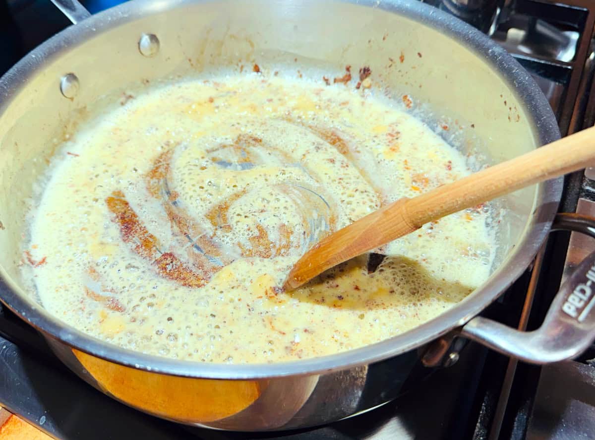 Pale yellow mixture of flour and butter being stirred with a wooden spoon in a deep sided steel saute pan.