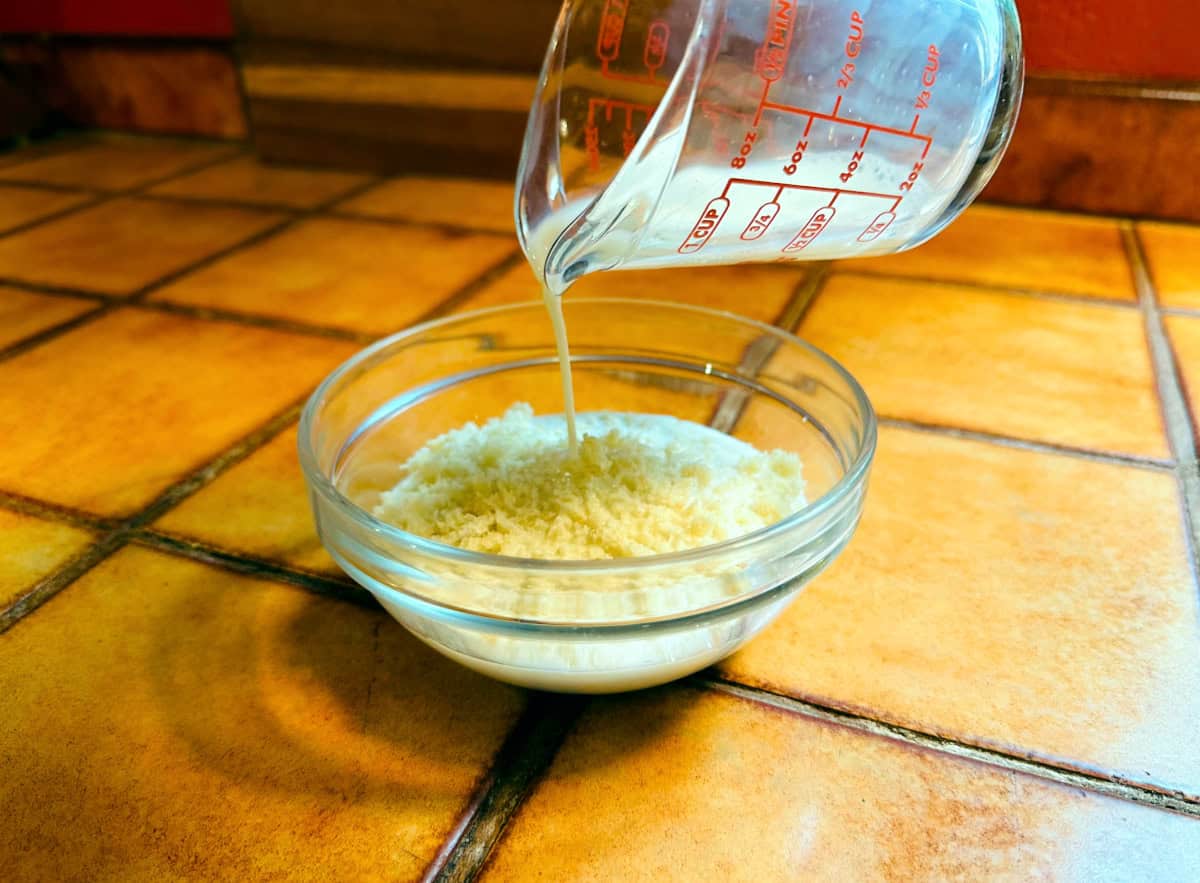 Milk being poured from a glass measuring cup into a small glass bowl of breadcrumbs.