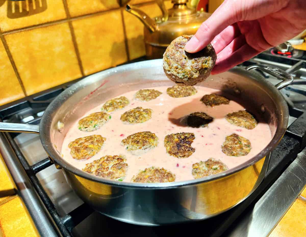 Cooked meatballs being added to pale pink sauce in a deep sided steel saute pan.