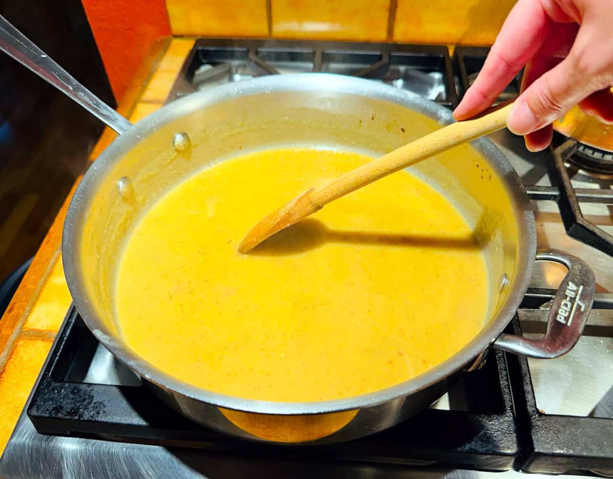 Chicken stock being stirred with a wooden spoon into flour and fat mixture in a deep sided steel saute pan.