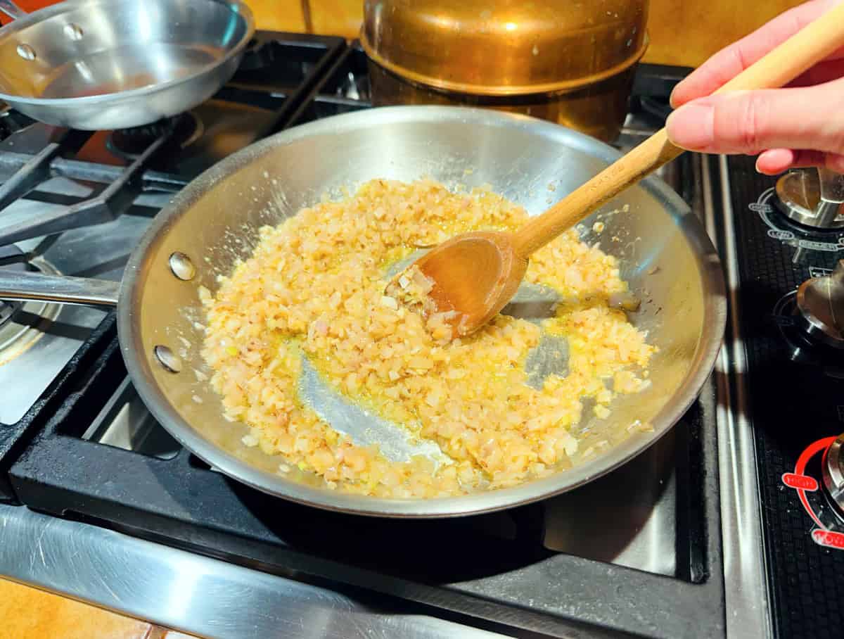 Shallots being stirred with a wooden spoon in a steel skillet on the stove.