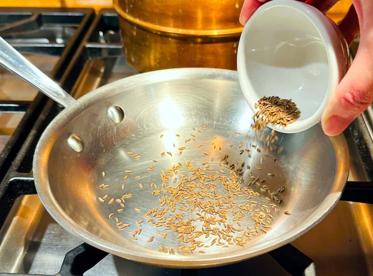 Small brown caraway seeds being tipped from a small white bowl into a small steel frying pan sitting on the stove.