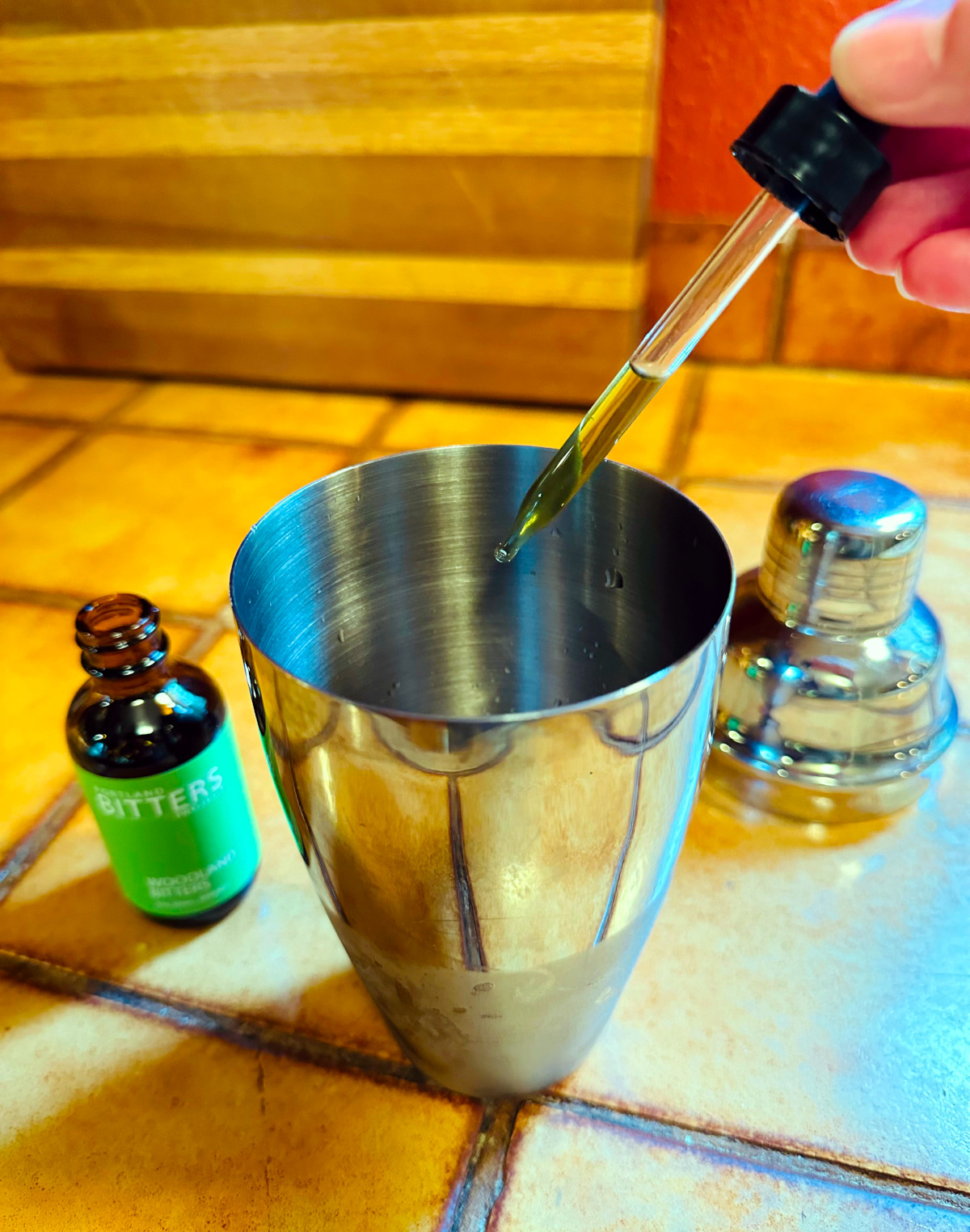 Dropper filled with amber colored liquid being held over a steel cocktail shaker next to a bottle of bitters.
