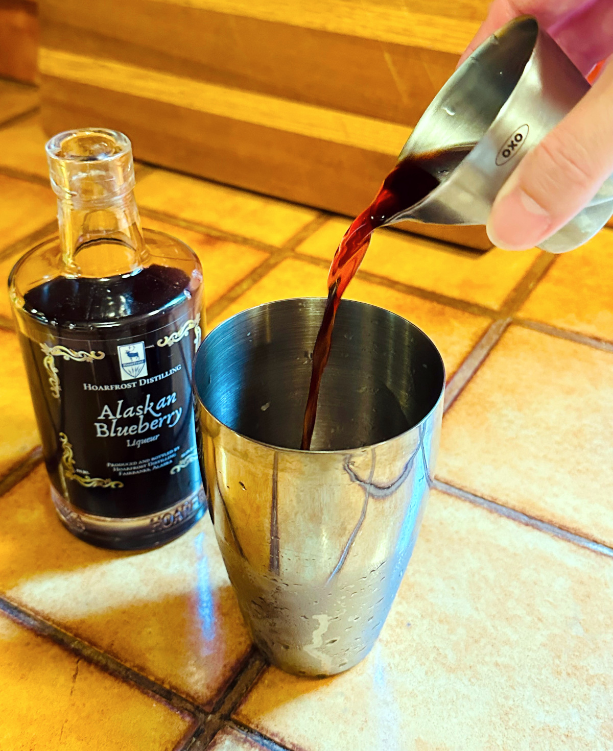 Dark violet colored liquid being poured from a steel measuring jigger into a steel cocktail shaker next to a bottle of blueberry liqueur.