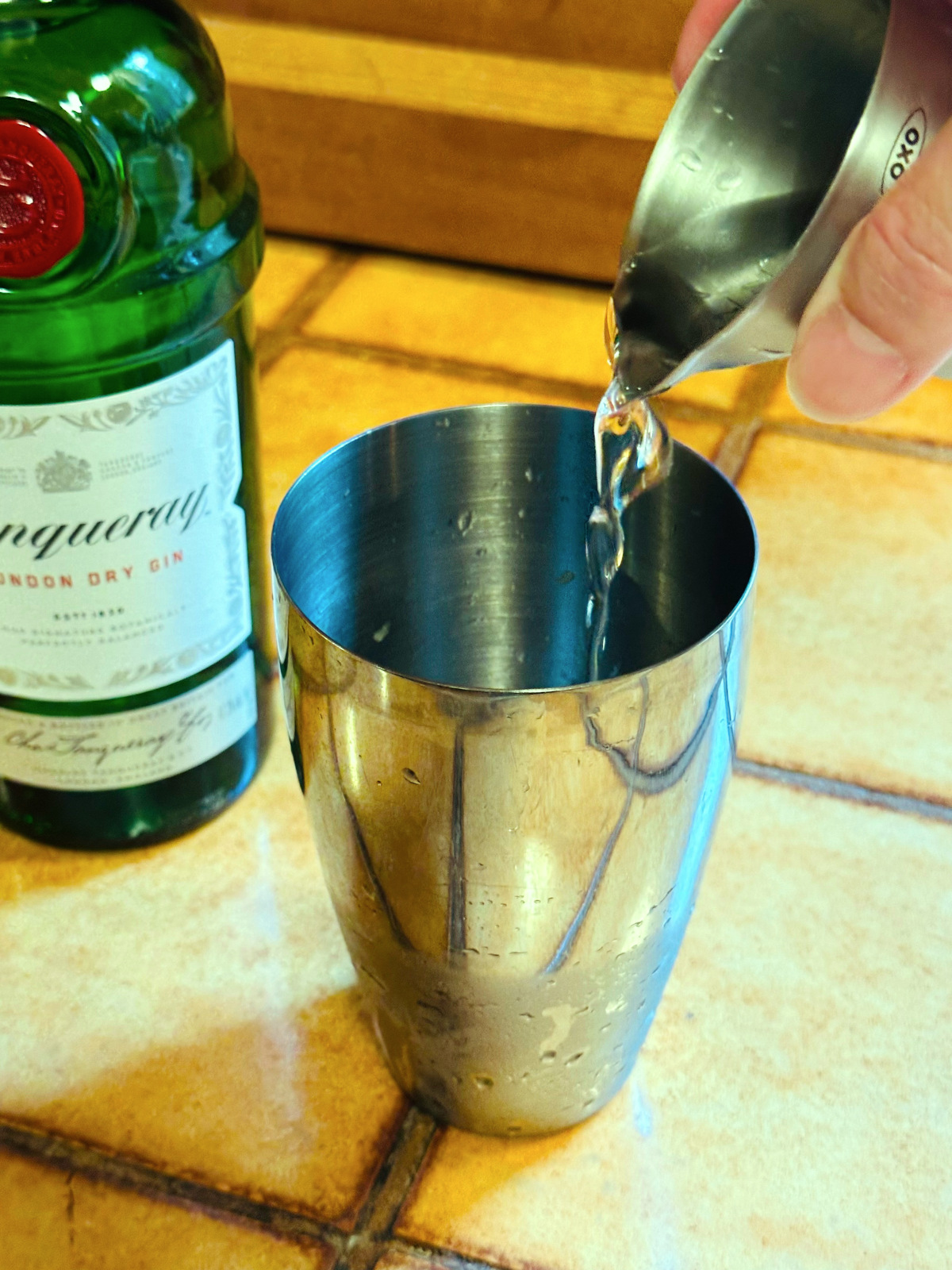 Clear liquid being poured from a steel measuring jigger into a steel cocktail shaker next to a bottle of gin.