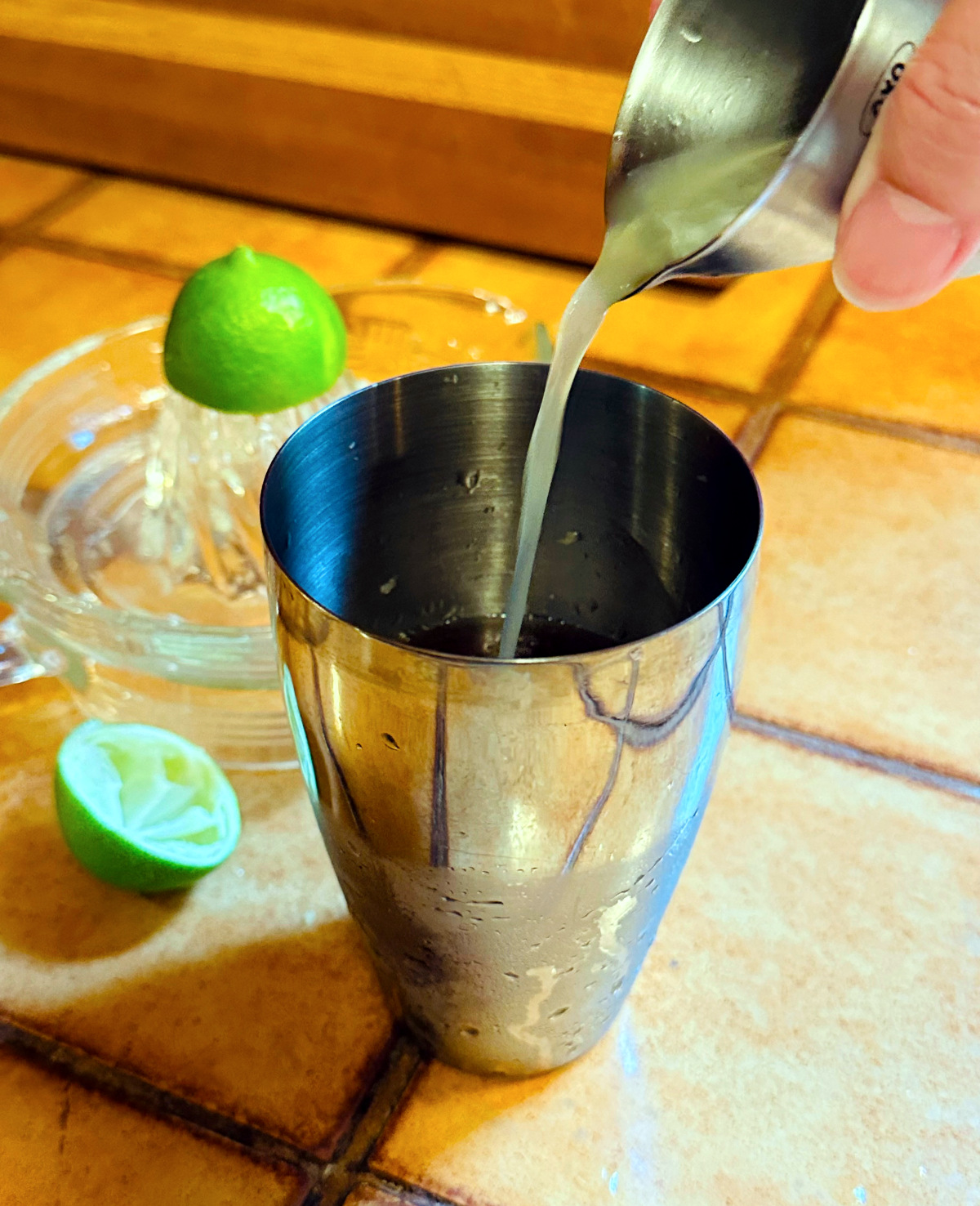Pale green liquid being poured from a steel measuring jigger into a steel cocktail shaker next to a glass citrus juicer and a couple lime halves.