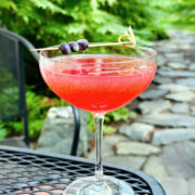 Cabin Club Cocktail (AKA Blueberry Pegu Club) in a coupe glass garnished with three blueberries skewered onto a cocktail pick.