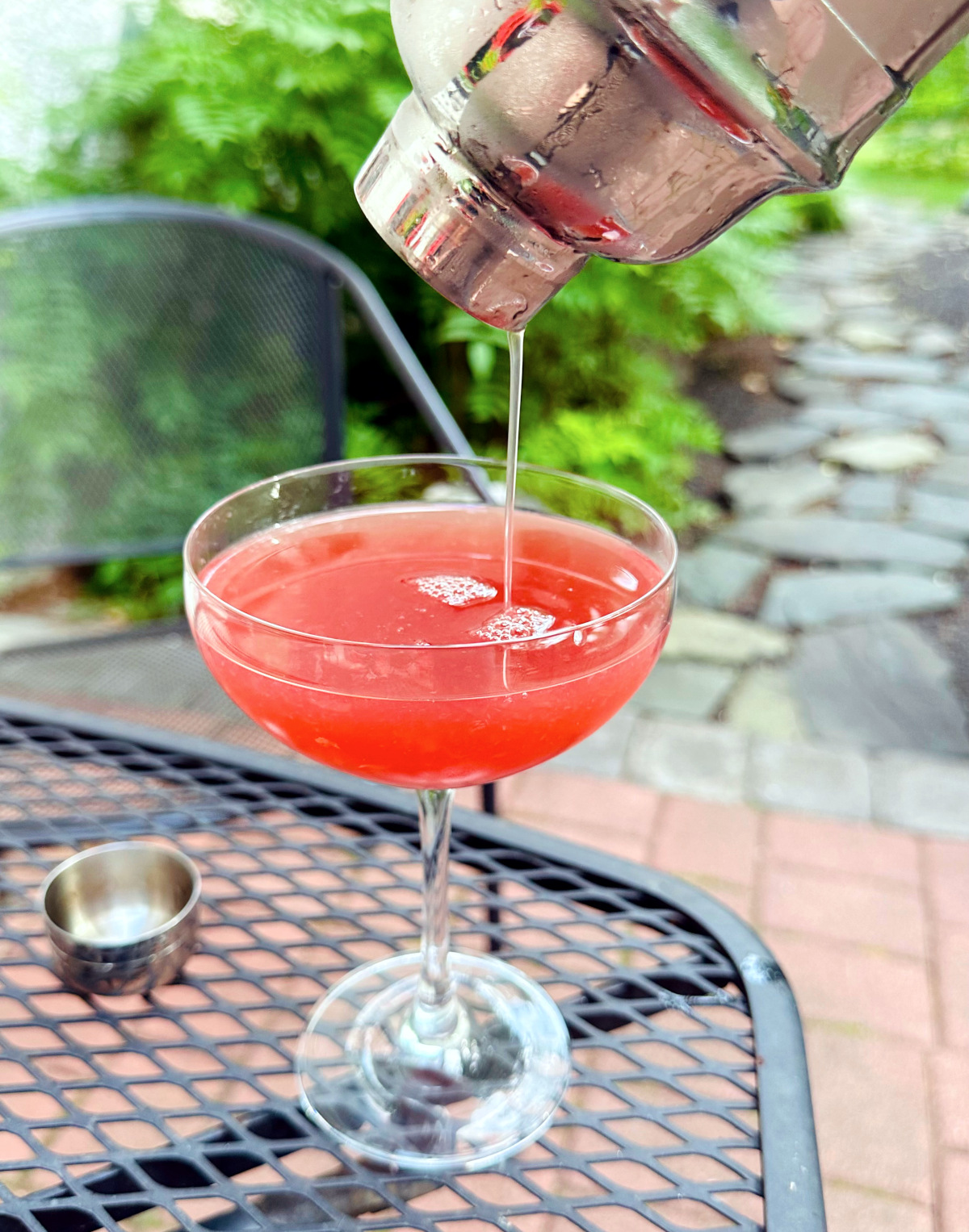 Dark pink liquid being poured from a steel cocktail shaker into a coupe glass sitting on a black metal outdoor table.