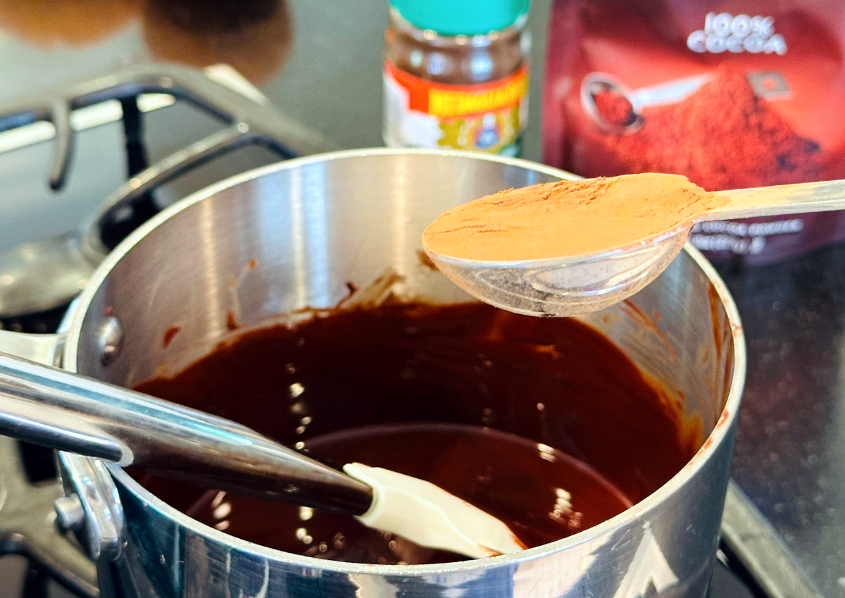 Cocoa in a steel measuring spoon being held over a small steel saucepan containing melted chocolate and a silicone spatula.