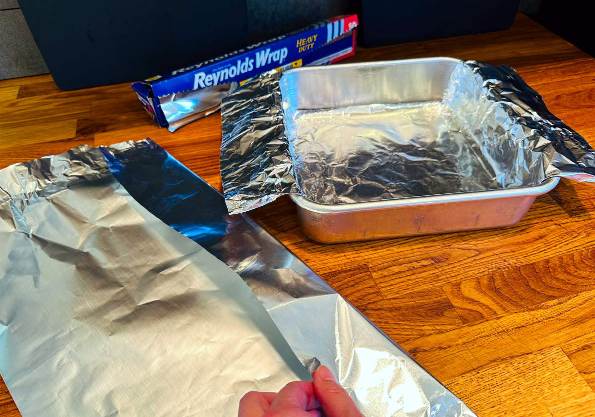 Foil being folded next to a square metal pan partially lined with foil.