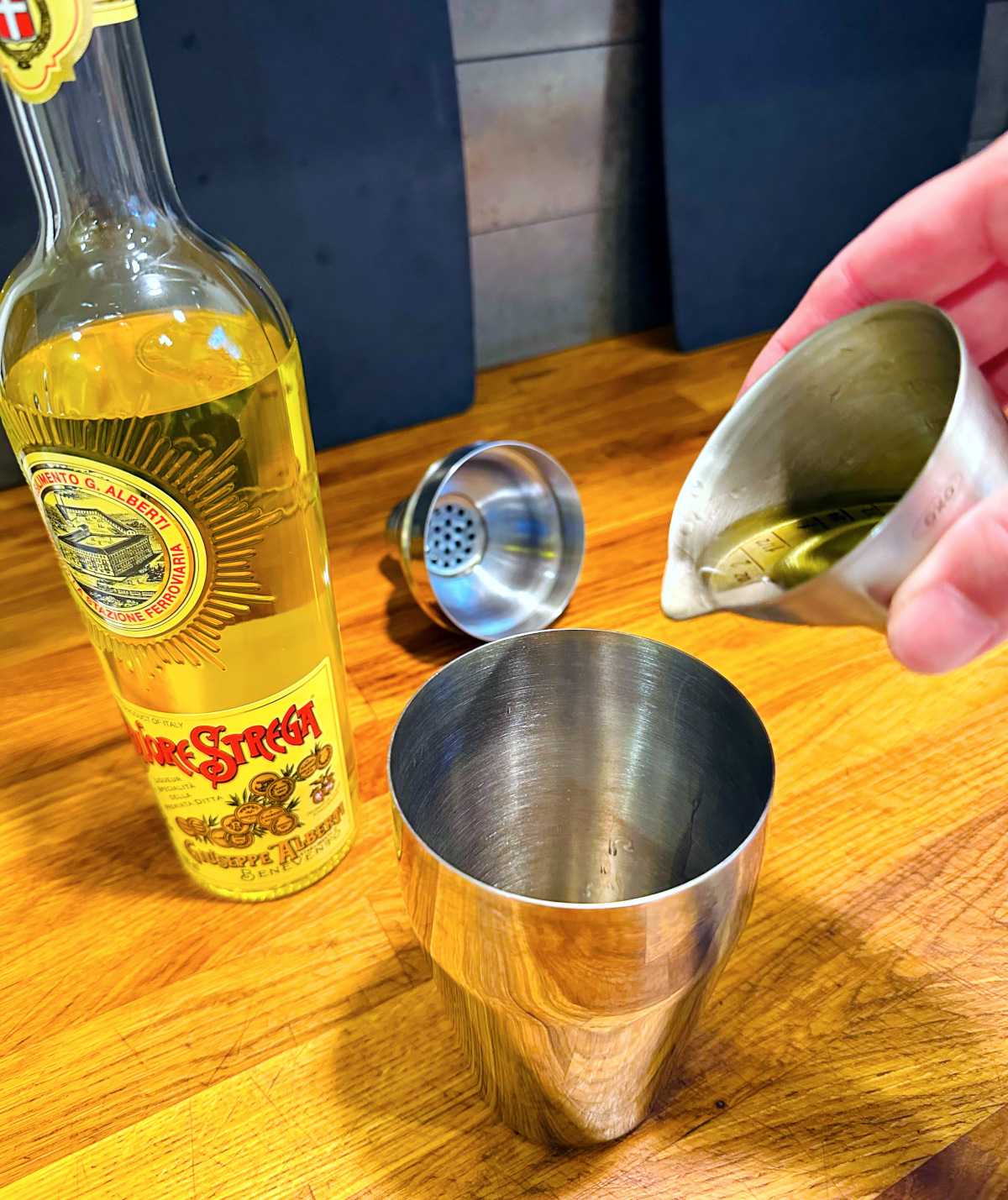 Yellow liquid being tipped from a steel measuring jigger into a cocktail shaker next to a bottle of Strega.