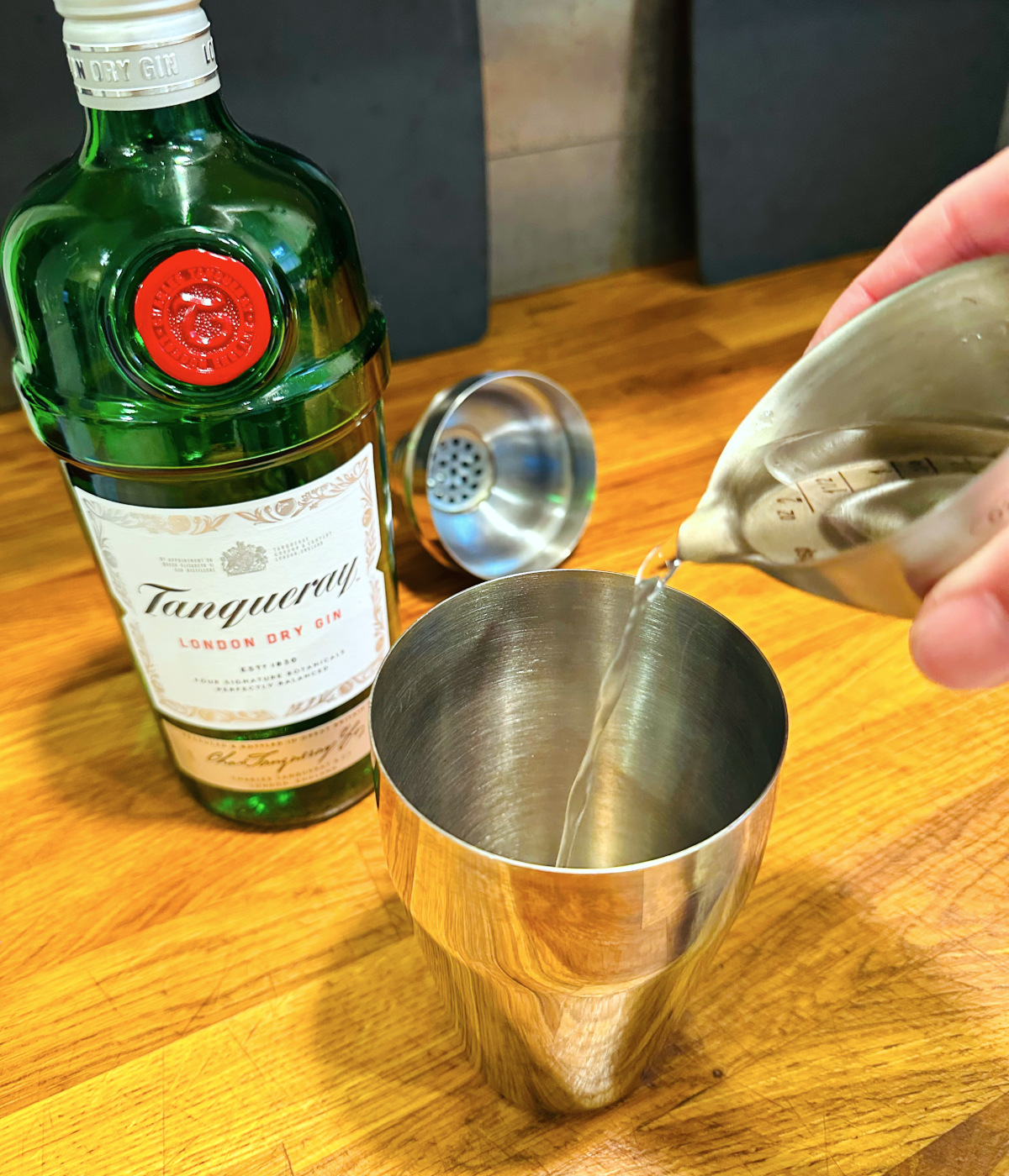 Clear liquid being tipped from a steel measuring jigger into a cocktail shaker next to a bottle of gin.