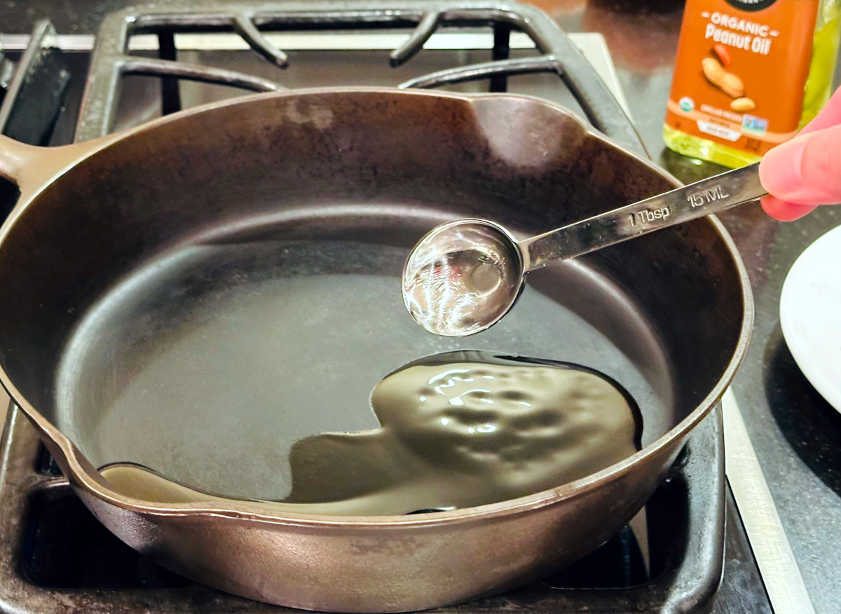 Pale yellow liquid being tipped from a steel measuring spoon into a cast iron skillet next to a bottle of peanut oil.