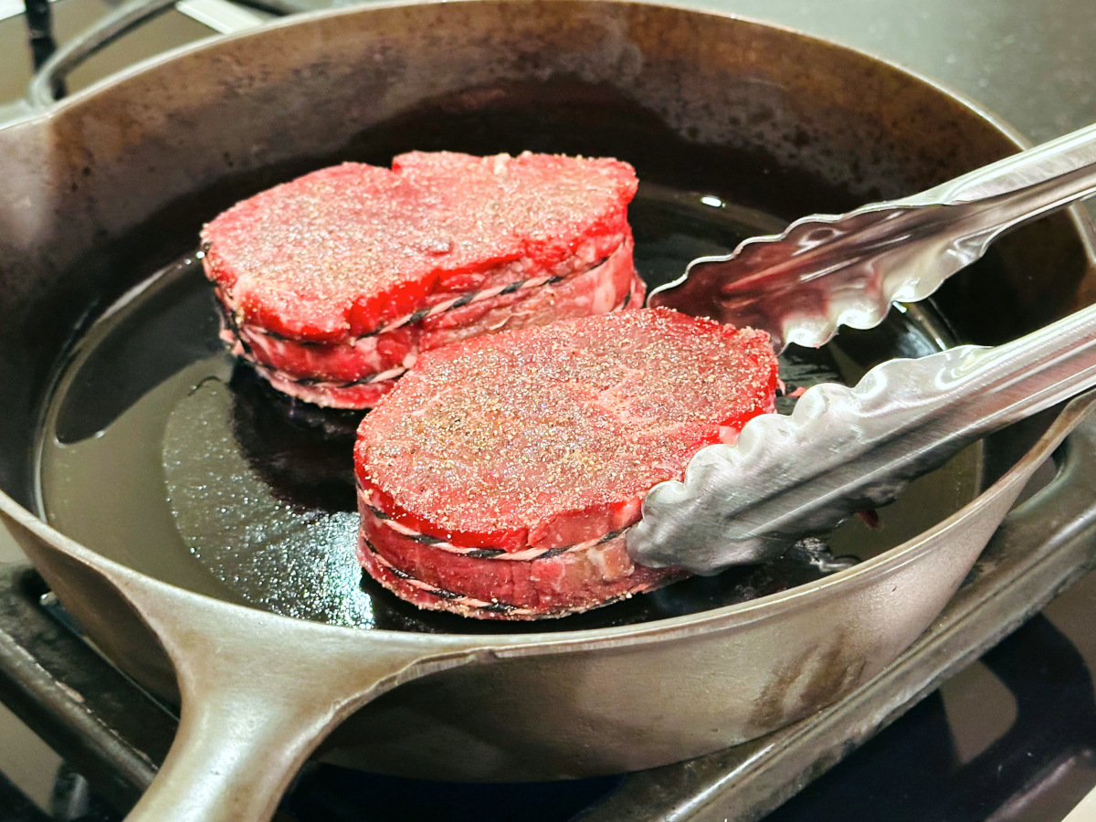 Two filet mignon steaks being placed with metal tongs into a cast iron skillet.