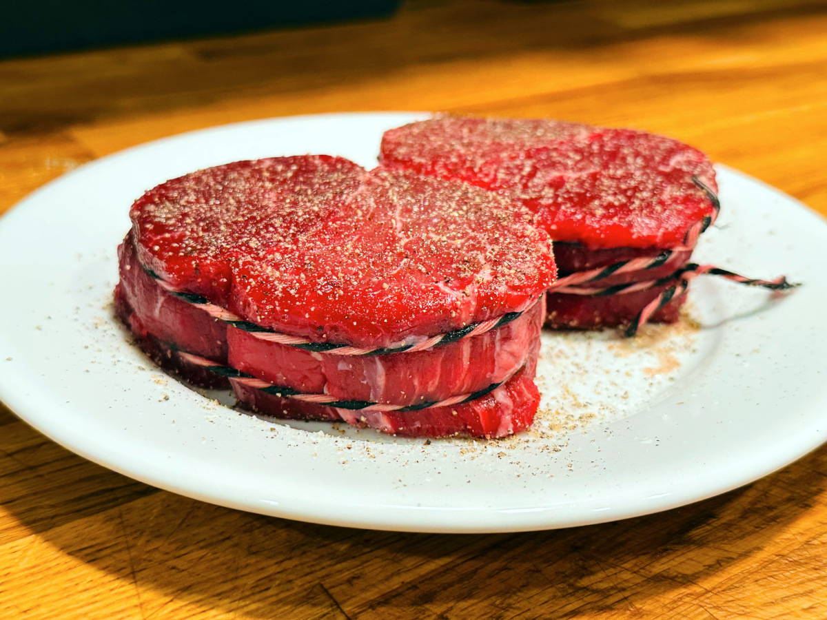 Two filet mignon steaks tied with string, seasoned with salt and pepper, and sitting on a white plate.
