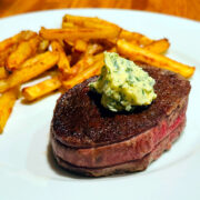 Pan seared filet mignon dolloped with compound butter and sitting on a white plate with some French fries.