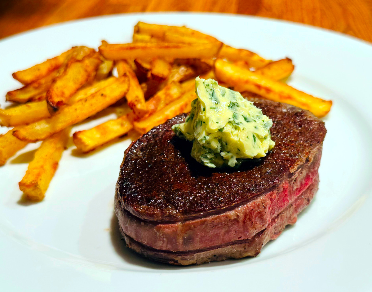 Pan seared filet mignon dolloped with compound butter and sitting on a white plate with some French fries.
