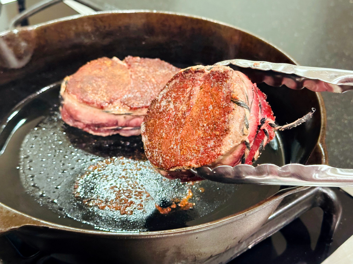 Steak in a cast iron skillet being flipped over with metal tongs.
