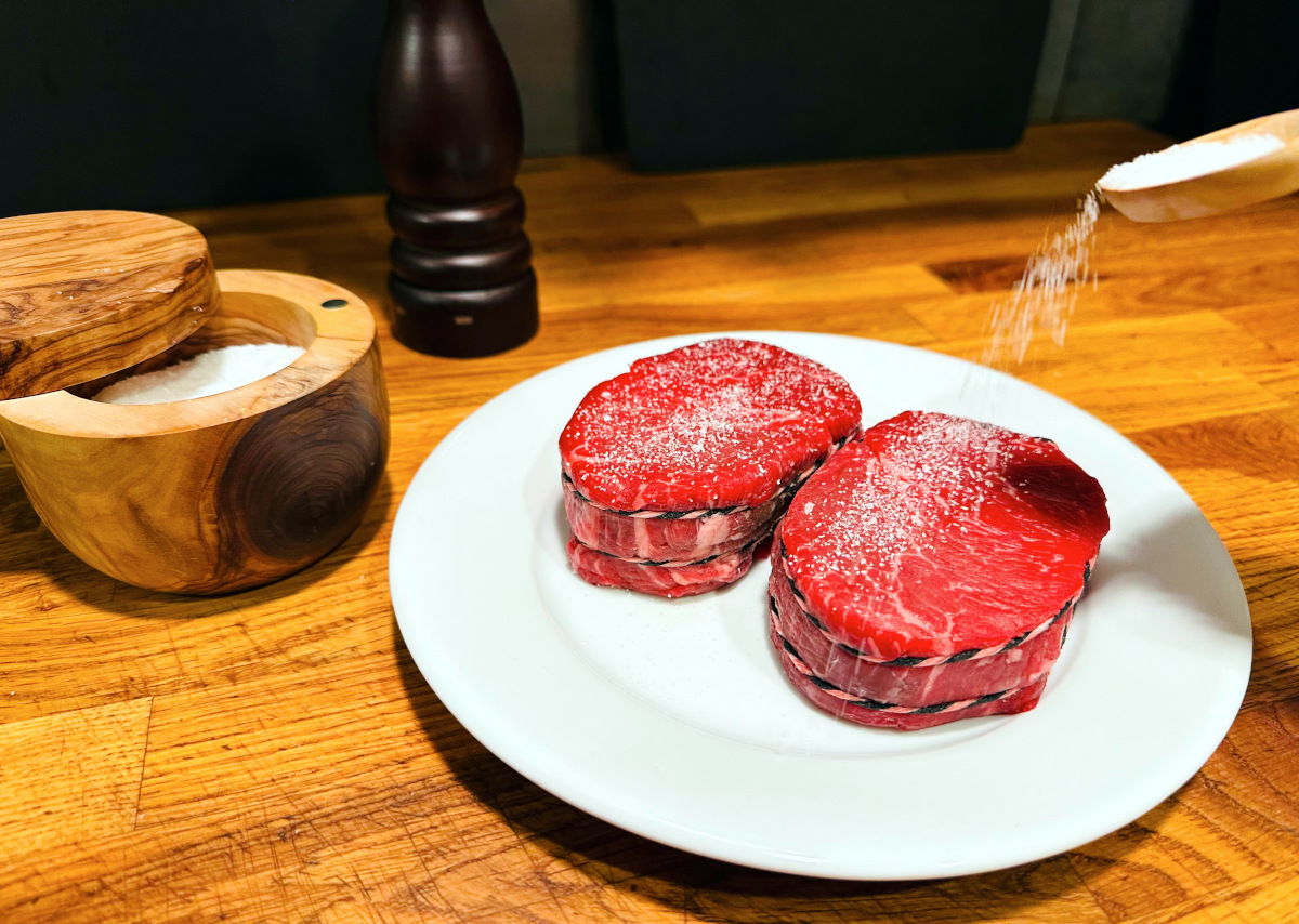 Salt being sprinkled from a small wooden spoon over two filet mignon steaks sitting on a white plate next to a salt cellar and a pepper grinder.