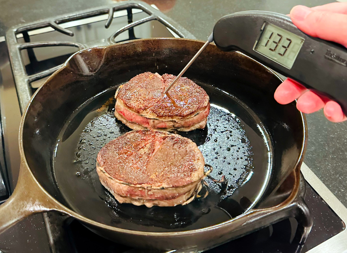 Steaks in a cast iron skillet being tested for temperature with a black digital thermometer.