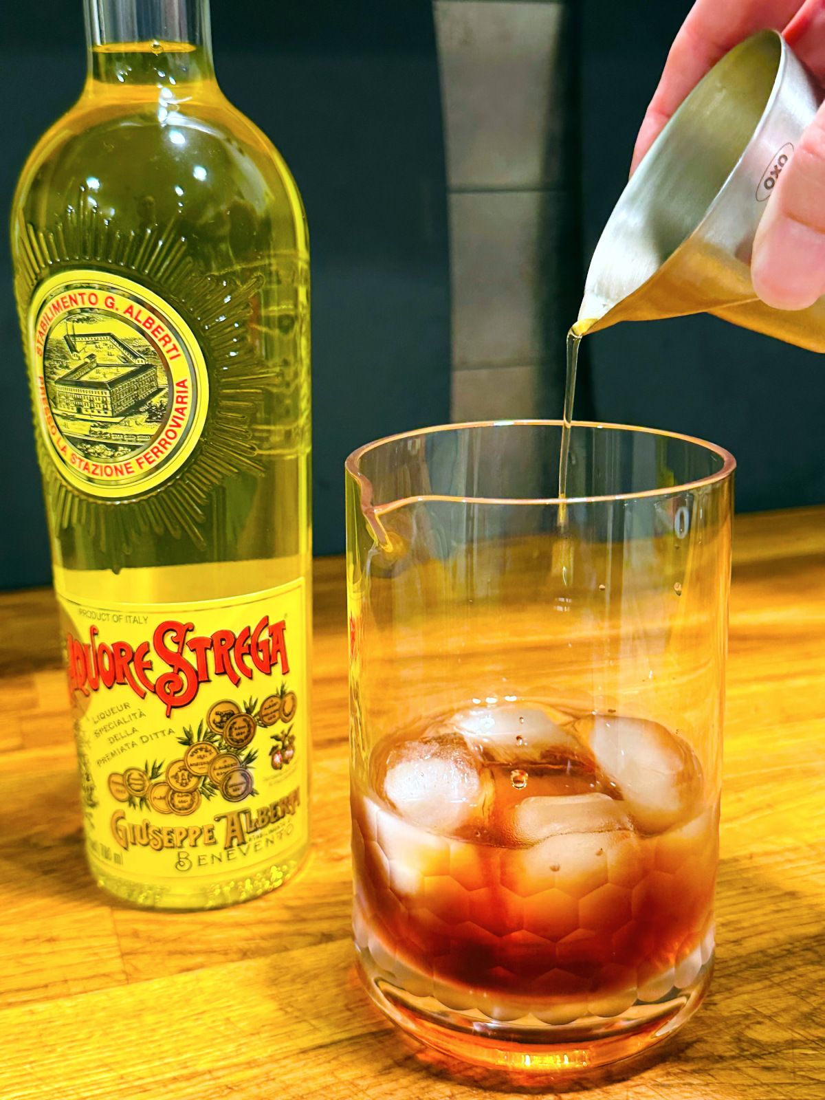 Yellow liquid being poured from a steel measuring jigger into a mixing glass with reddish liquid and ice next to a bottle of Strega.