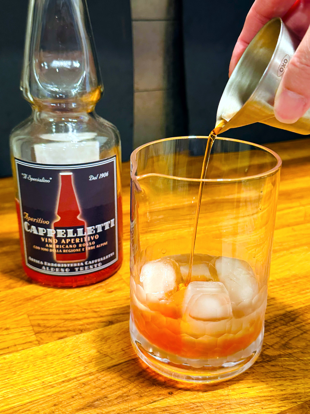 Pale red liquid being poured from a steel measuring jigger into a mixing glass with ice next to a bottle of Cappelletti.