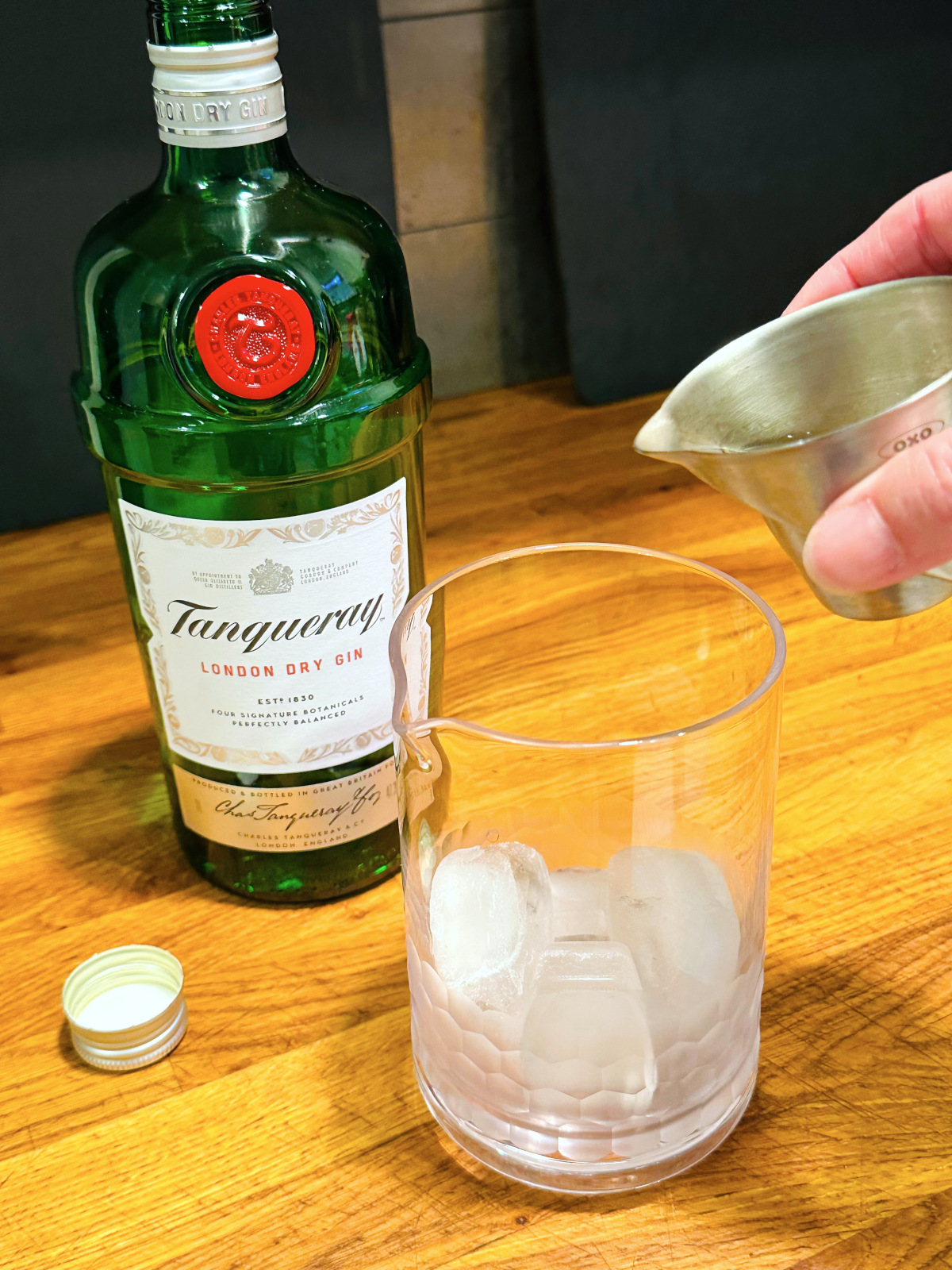 Steel measuring jigger being held over a mixing glass with ice next to a bottle of Tanqueray gin.