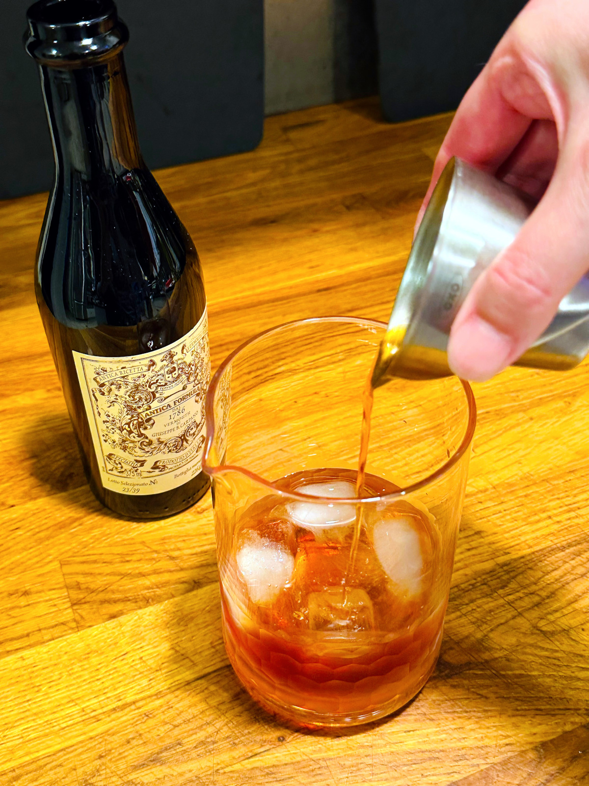 Reddish liquid being poured from a steel measuring jigger into a mixing glass with ice next to a bottle of sweet vermouth.