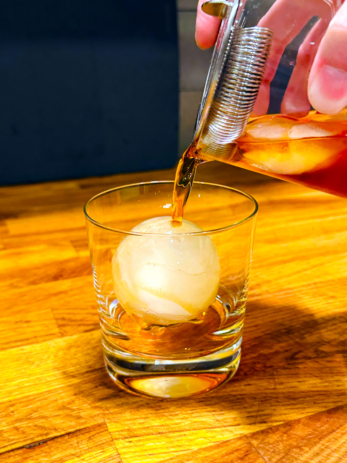 Dark reddish liquid being strained from a mixing glass over a large sphere of ice in an old fashioned glass.