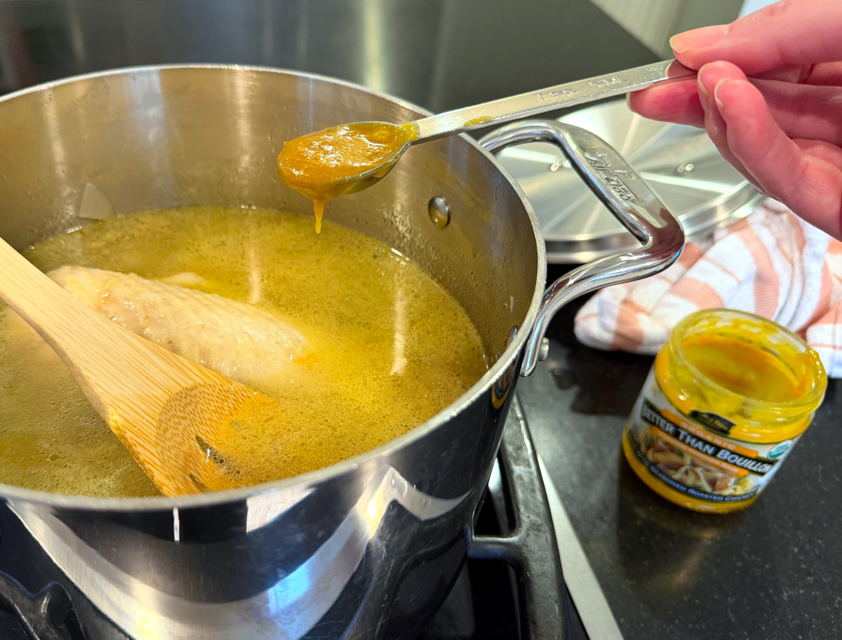 Dark yellow paste being tipped form a steel measuring spoon into yellow liquid in a steel saucepan next to a jar of bouillon paste.