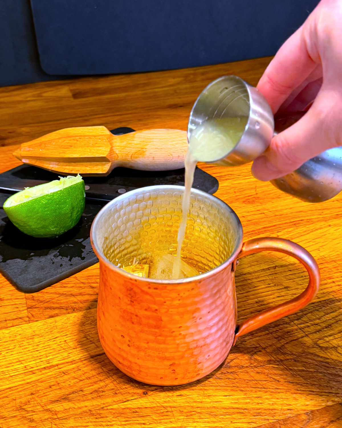 Pale green liquid being tipped from a steel measuring jigger into a copper mug next to half a lime and a reamer sitting on a small cutting board.