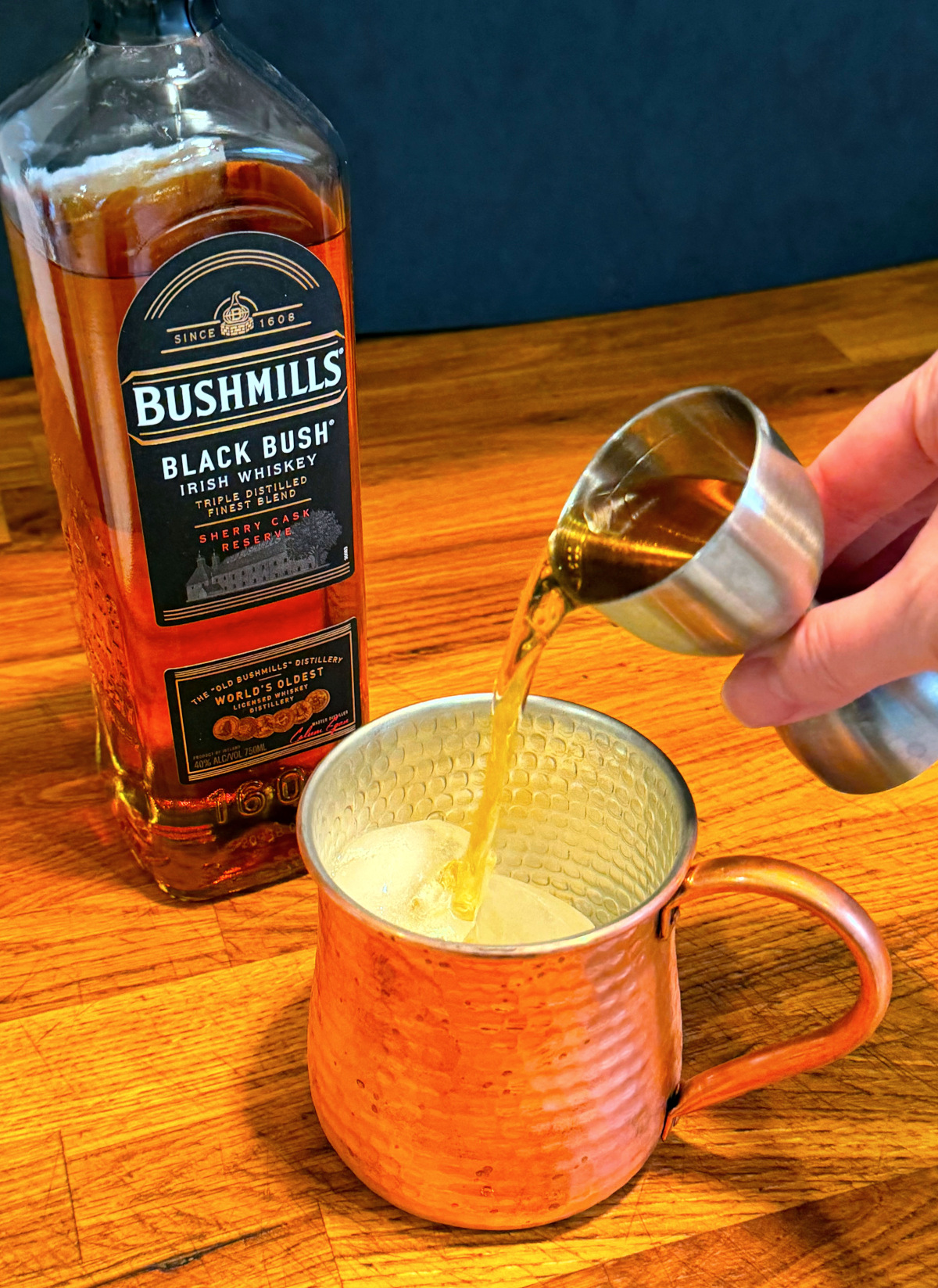 Amber liquid being tipped from a steel measuring jigger into a copper mug next to a bottle of Irish Whiskey.