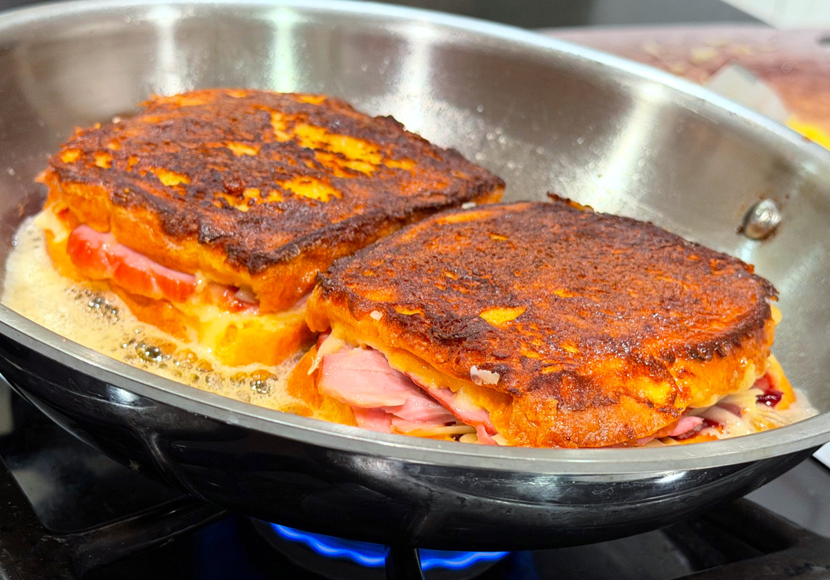 Two golden brown sandwiches frying in butter in a large steel frying pan.