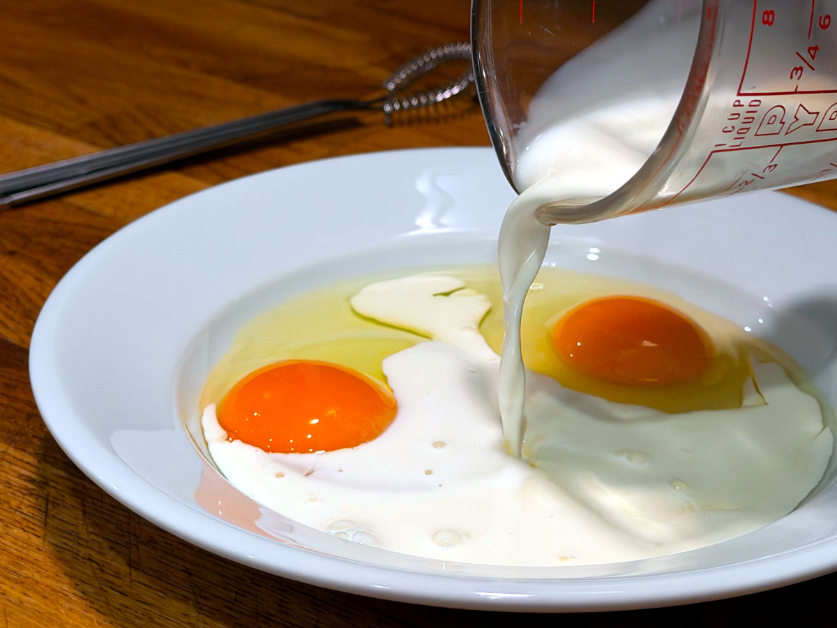 Milk being poured from a glass measuring cup into a shallow white bowl containing two cracked raw eggs.