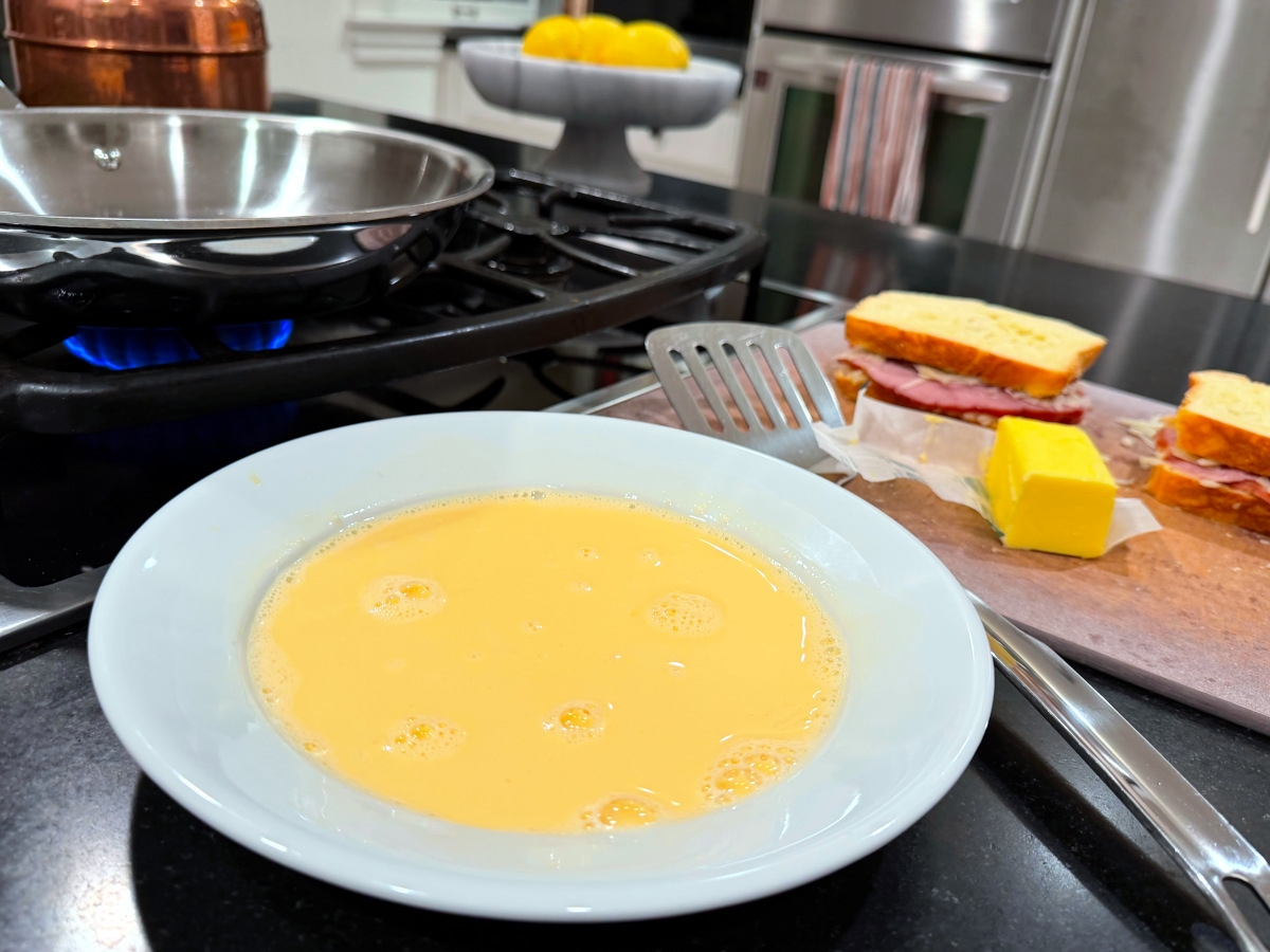 Beaten egg mixture in a shallow white bowl sitting between a large steel frying pan heating on the stove and a cutting board holding two sandwiches, a block of butter, and a metal spatula.