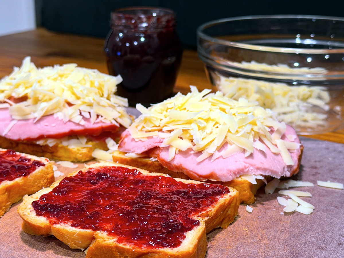 Two slices of bread topped with ham and shredded cheese sitting next to two slices of bread spread with dark red jam.