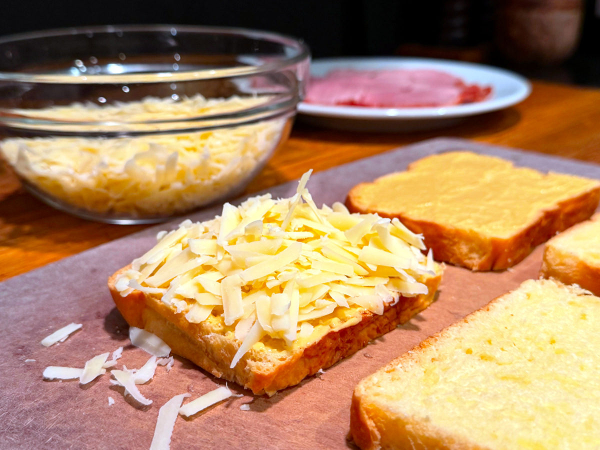 Slice of bread sprinkled with shredded cheese next to a slice of bread covered in mustard on a cutting board in front of a bowl of shredded cheese and a plate of ham.