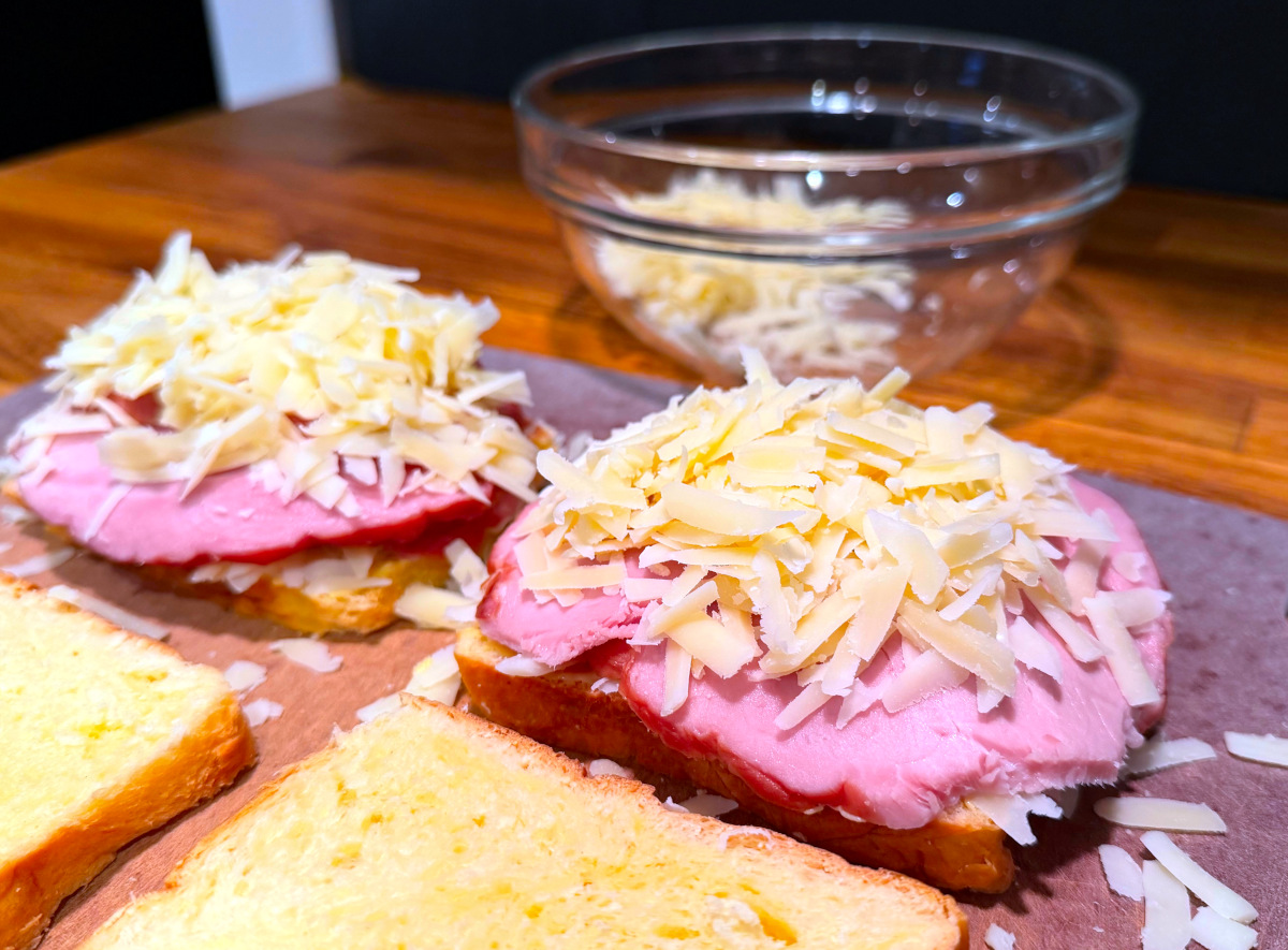 Two slices of bread topped with ham and shredded cheese in front of a glass bowl containing shredded cheese.