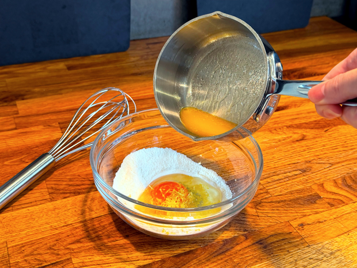 Melted butter being tipped from a small steel saucepan into a small glass bowl containing dry ingredients, egg, and lemon zest.