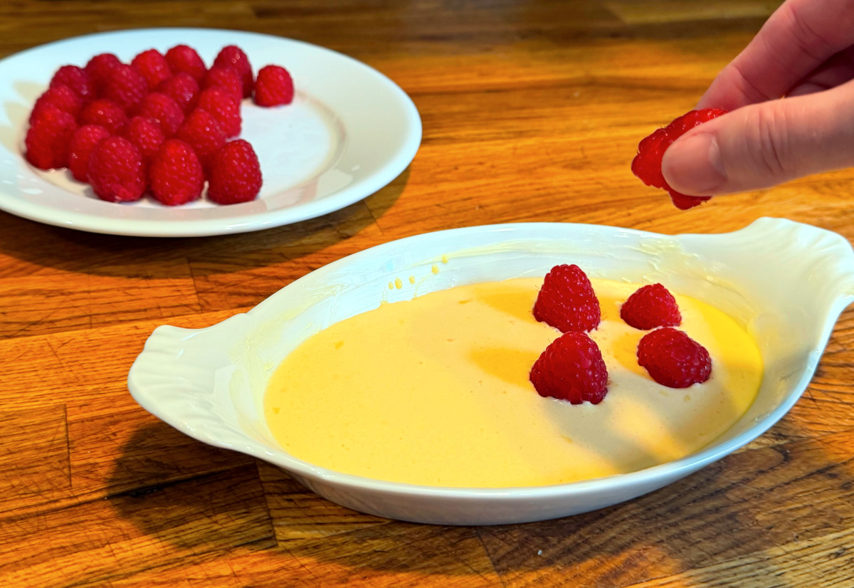 Fresh raspberries being dotted across yellow batter in a small white porcelain baking dish next to a plate full of fresh raspberries.