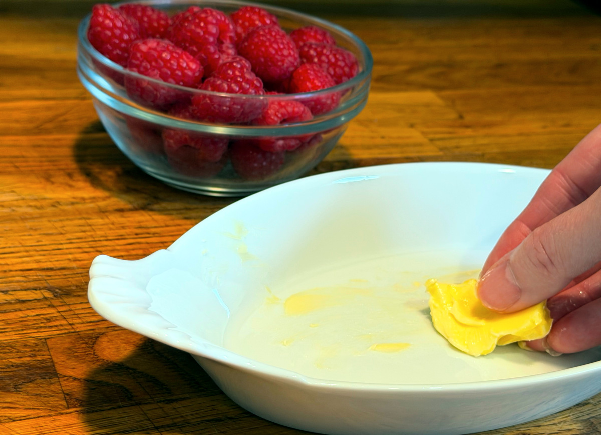 Small white oval baking dish being greased with butter next to a small bowl of fresh raspberries.