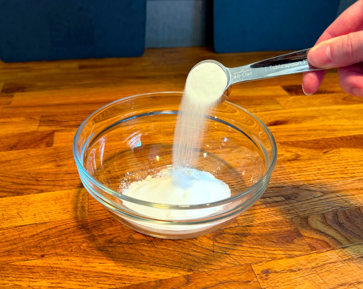 Tipping sugar from steel measuring spoon into small glass bowl containing flour.