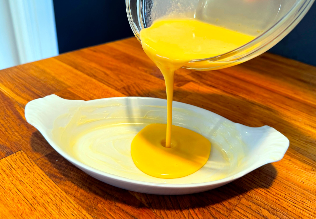 Yellow batter being poured from a glass bowl into a small white oval baking dish greased with butter.