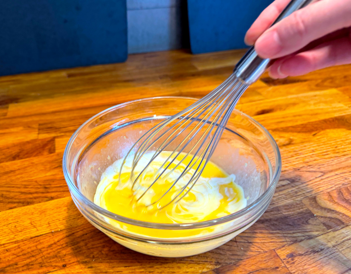 Milk being whisked into yellow batter in a small glass bowl.