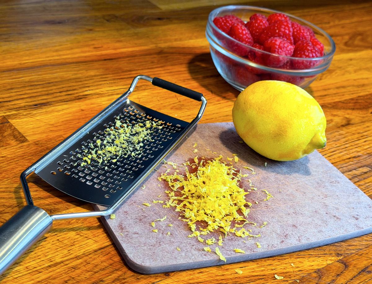 Lemon zest on a cutting board between a microplane zester and a lemon sitting in front of a small bowl of fresh raspberries.