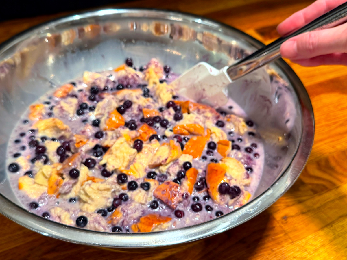 Blueberries being stirred into bread cubes and custard mixture in a large steel bowl.