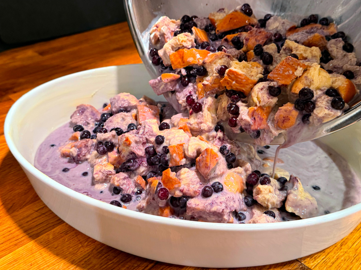Pale purple mixture of wet bread cubes, custard liquid, and blueberries being tipped from a steel bowl into a white oval baking dish.