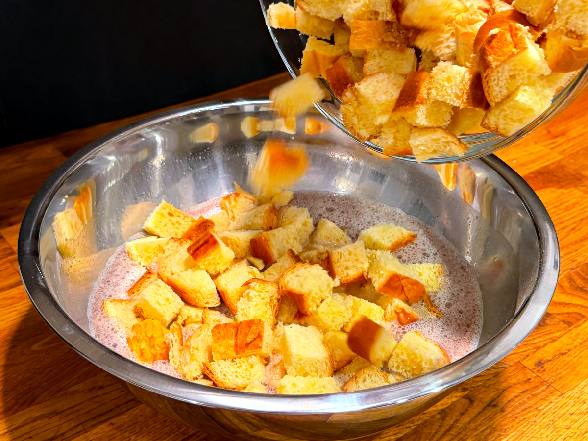 Bread cubes being tipped from a glass bowl into tan liquid in a large steel bowl.