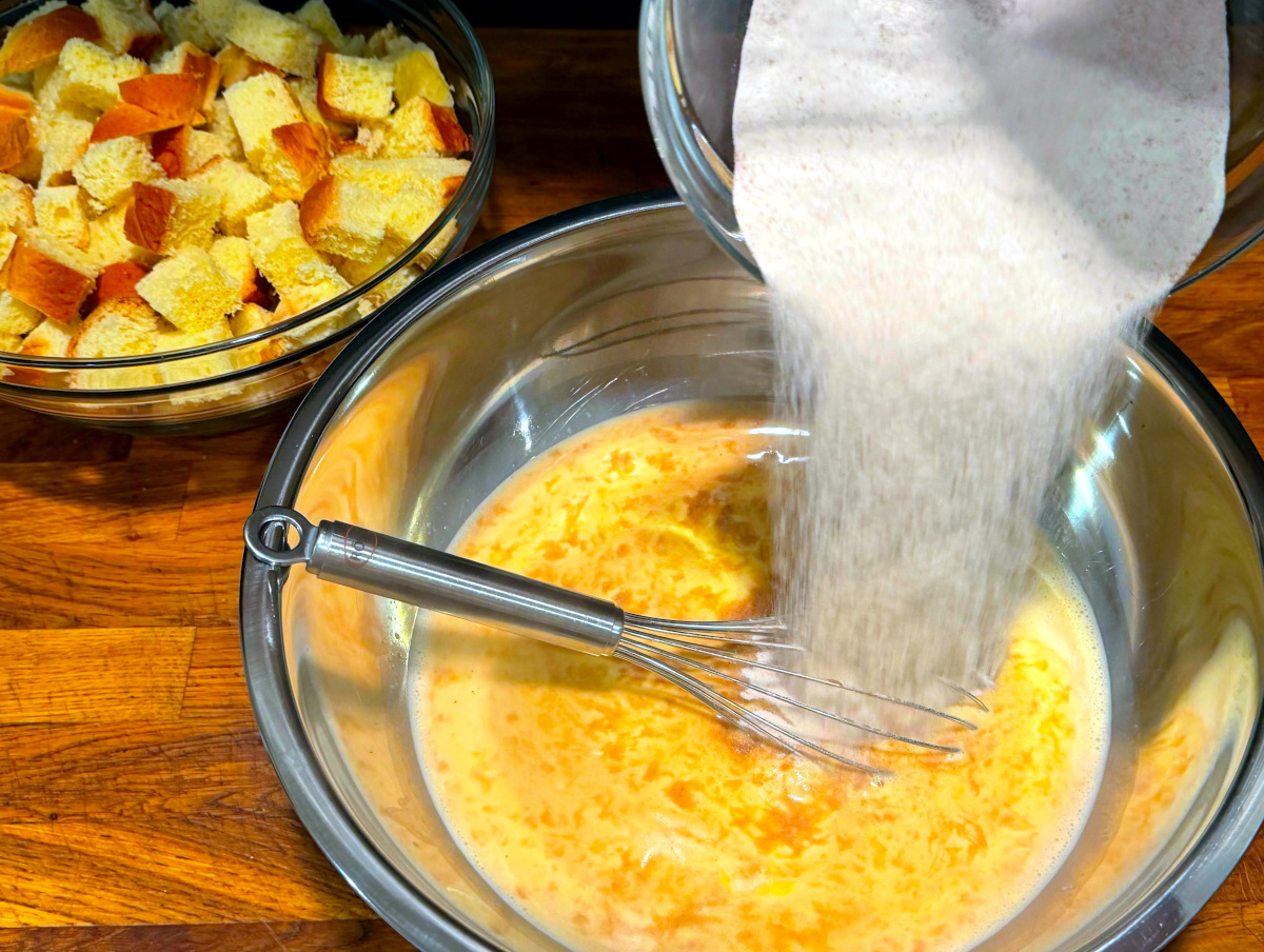 Sugar being tipped from a glass bowl into yellow liquid swirled with vanilla extract in a large steel bowl with a whisk sitting inside next to a glass bowl of bread cubes.
